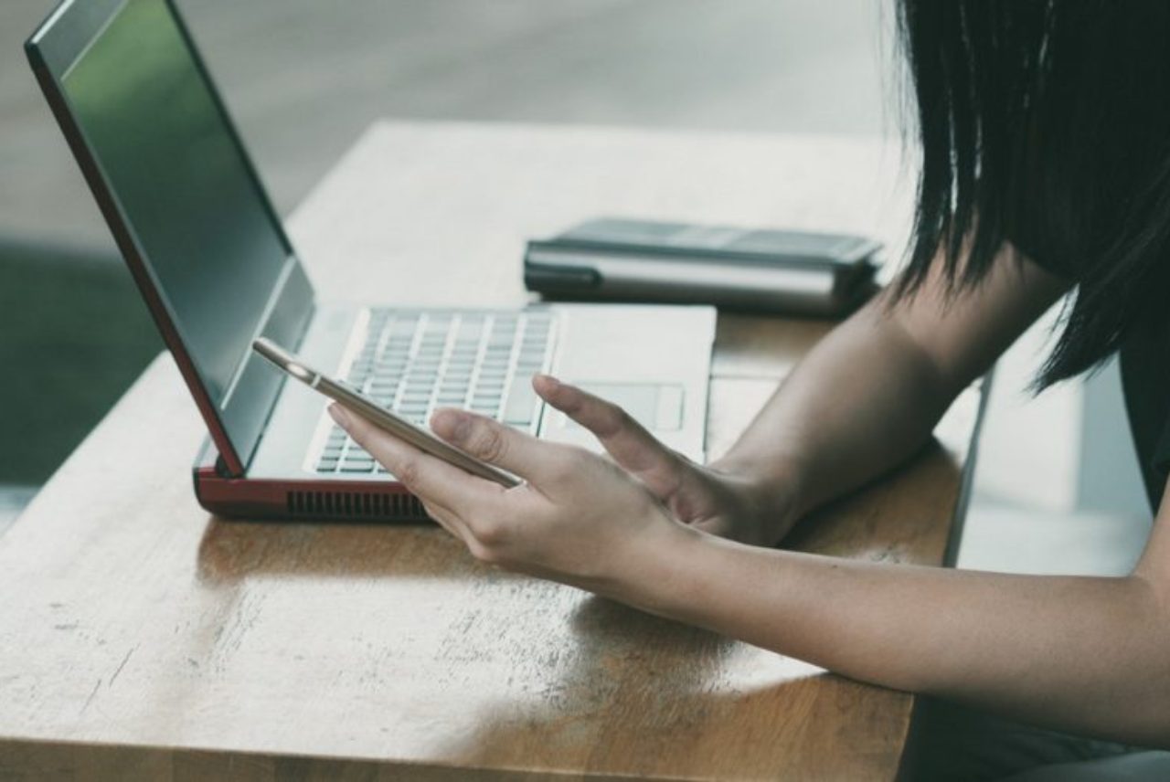 female-arms-leaning-on-table-holding-smartphone-with-open-laptop-on-table